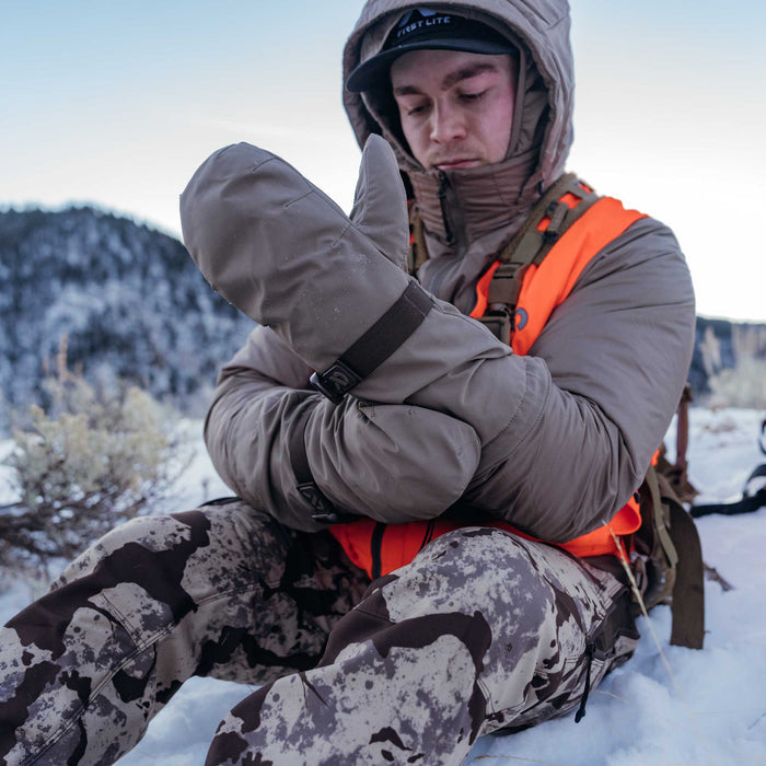 Grey-taupe insulated mittens with wrist straps and buckles are worn by a man sitting in snow.