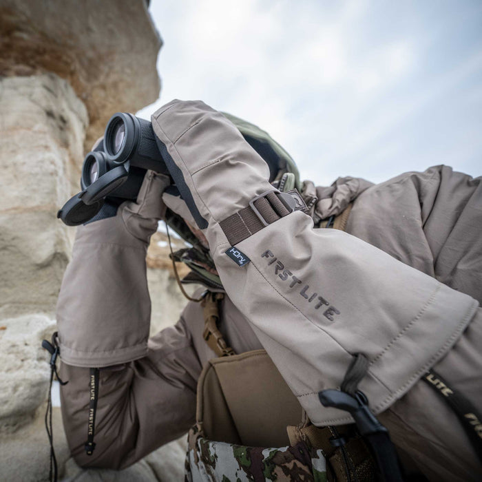 Tan First Lite HDry oversized mitts with an adjustable wrist strap worn by a person looking through binoculars.