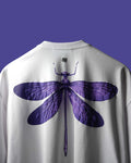 A close-up of the back of a white crewneck sweatshirt featuring a large purple dragonfly graphic print, set against a solid purple background.