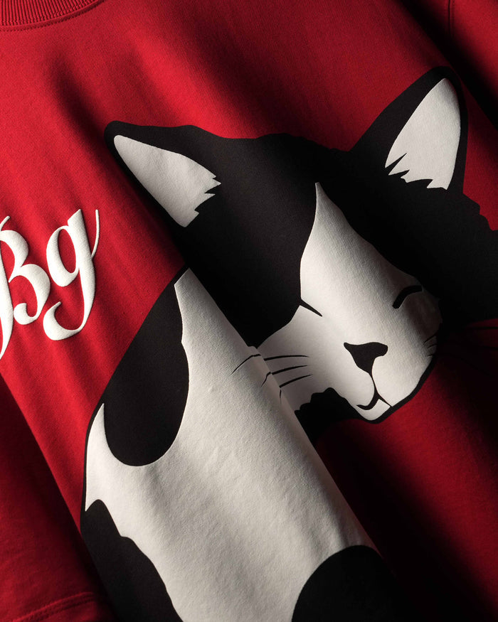 A close-up of a red crewneck sweatshirt featuring a large black and white cat graphic and partial white script text on the chest.