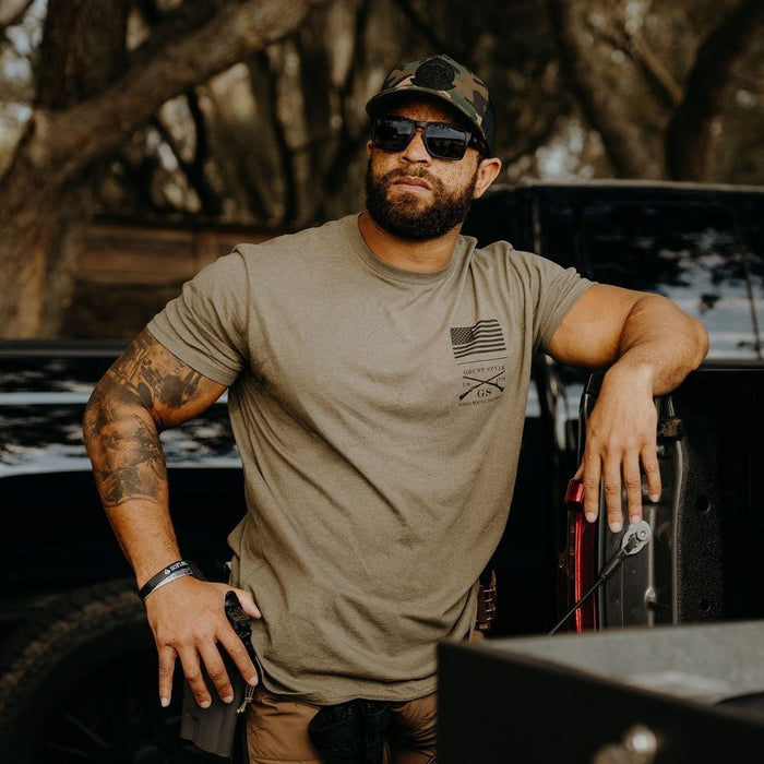 Man wearing a camouflage cap and sunglasses, leaning against a vehicle with a forest background.