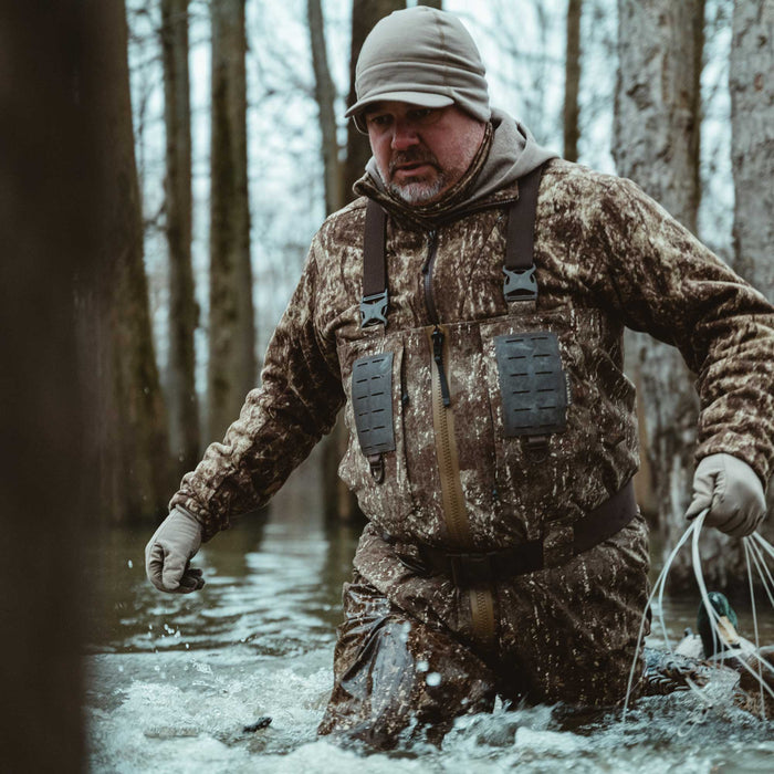 Brown camouflage wader suit with suspenders, a front zipper, and chest utility panels, shown on a man walking through water.