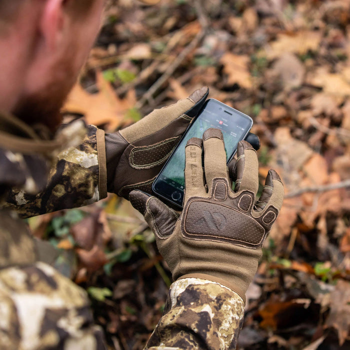 Tan and brown tactical gloves with perforated leather knuckle pads and reinforced palms worn by a person using a smartphone outdoors.