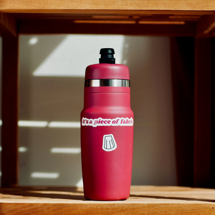 Red water bottle with 'it's a piece of fabric' and '[skirt outline]' stickers, on a wooden shelf.