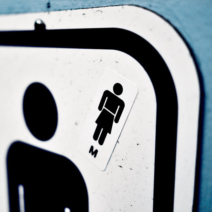The top corner of a men's washroom sign, with a small sticker over top (above shoulder), of a men's restroom symbol, only with a skirt instead of pants, and an 'M'.