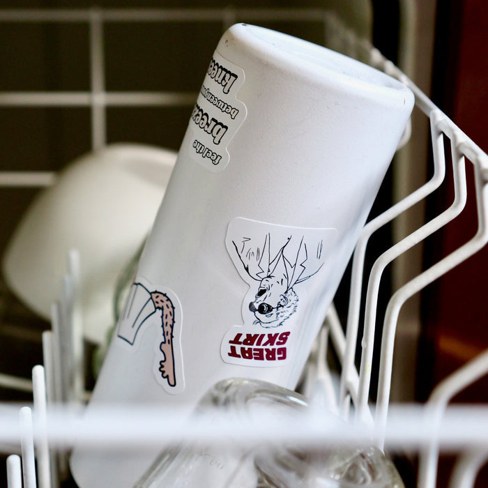 A white to-go mug with stickers upside down in the top rack of a dishwasher.
