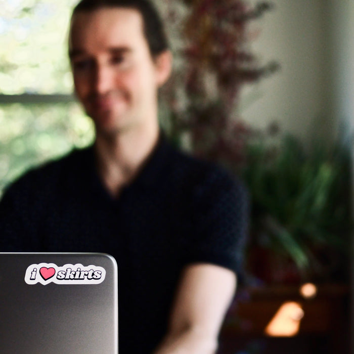 Male human working on a laptop, with 'i <3 skirts' sticker in focus on top corner of laptop lid.
