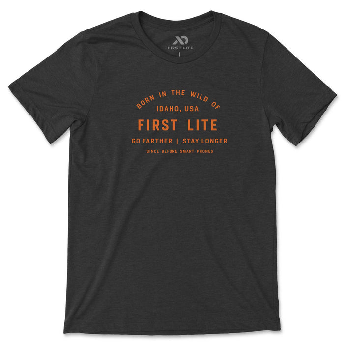 A heather charcoal t-shirt featuring orange text "Born in the Wild of Idaho, USA" and "First Lite - Go Farther | Stay Longer", with a small First Lite logo on the collar.