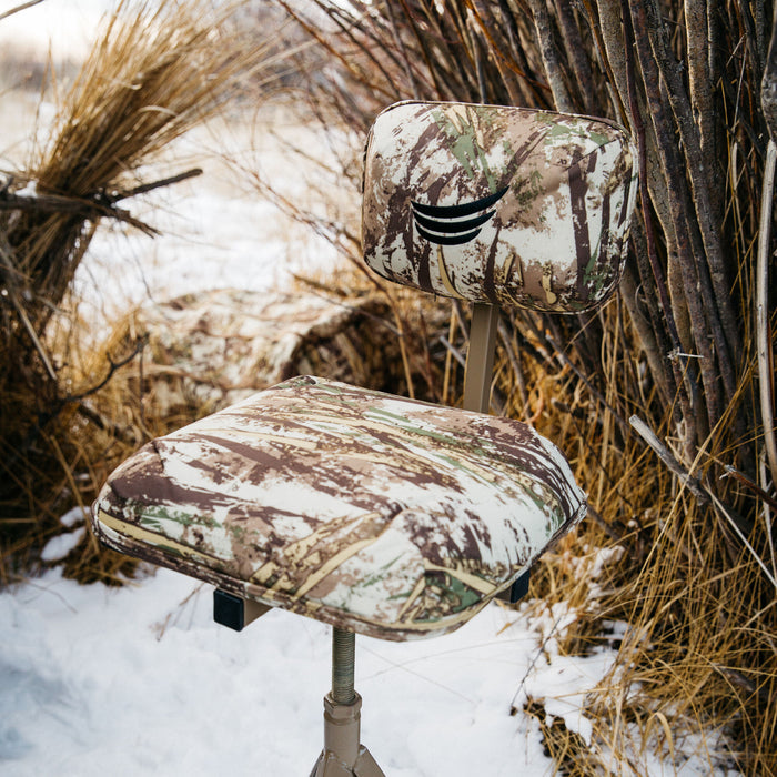 Camouflage hunting chair with an adjustable metal base and a black logo on the backrest, set in a snowy, brushy landscape.