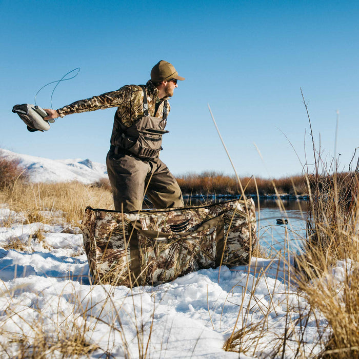 A camouflage layout blind with an open top rests on snowy ground near a river, beside a hunter throwing a duck decoy.
