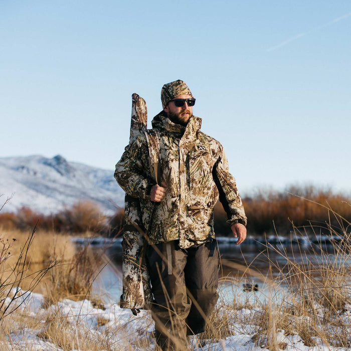 A man wearing a wetlands camouflage hunting jacket, matching hat, and dark waders carries a matching gun case through a snowy marsh.