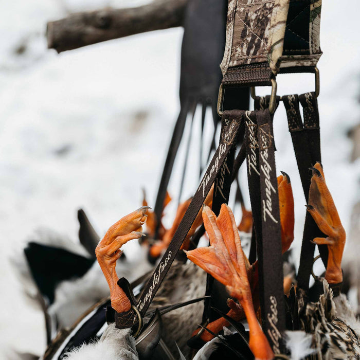 Camouflage game strap with brass hardware holding several waterfowl by their bright orange webbed feet on brown straps branded "Tanglefree" against a snowy background.