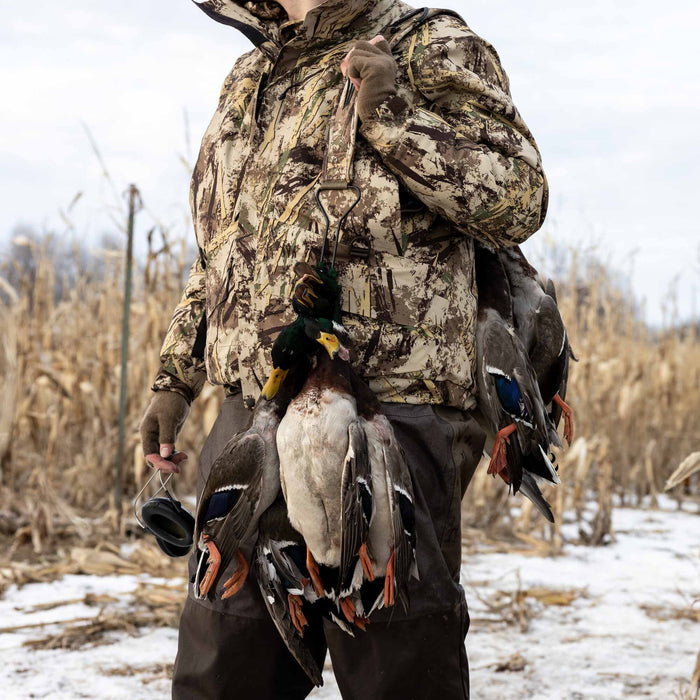 A hunter wears a multi-tone wetlands camouflage hooded jacket with brown fingerless gloves, carrying ducks in a snowy field.