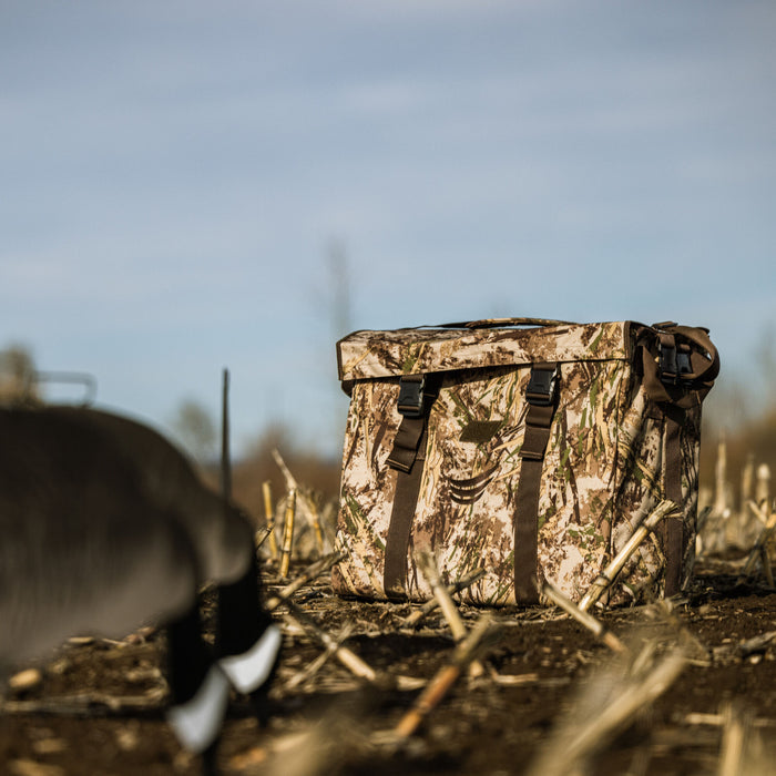 A camouflage waterfowl bag with a roll-top closure and two front buckle straps sits in a stubble field next to a goose decoy.