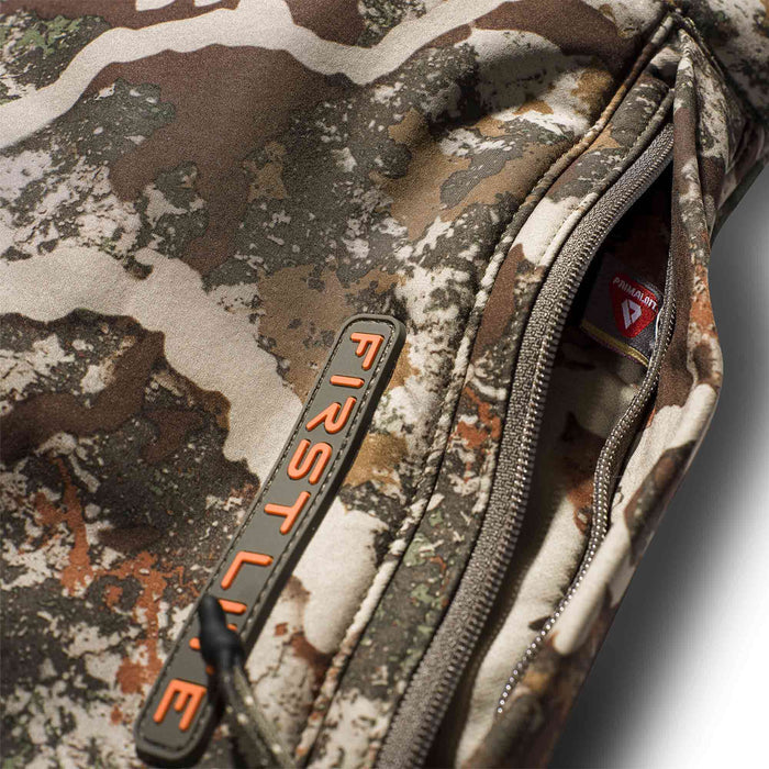 Close-up of a camouflage jacket with an open zippered pocket showing a Primaloft tag and an orange First Lite zipper pull.