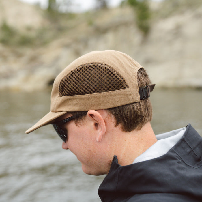 A tan 5-panel cap featuring dark mesh ventilation panels and a black adjustable strap, worn by a person.