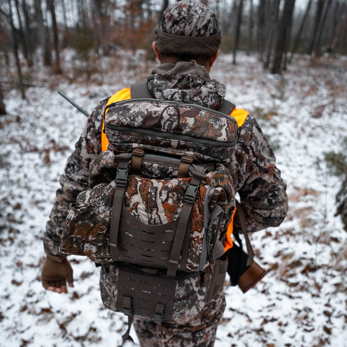 A multi-tone camouflage hunting backpack with multiple compartments and a laser-cut panel, worn by a hunter in a snowy woods.