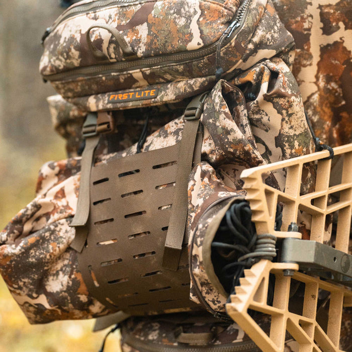 A First Lite Fusion camouflage hunting backpack featuring the brand logo, an olive green MOLLE panel, and a tan tree stand attachment on its side.