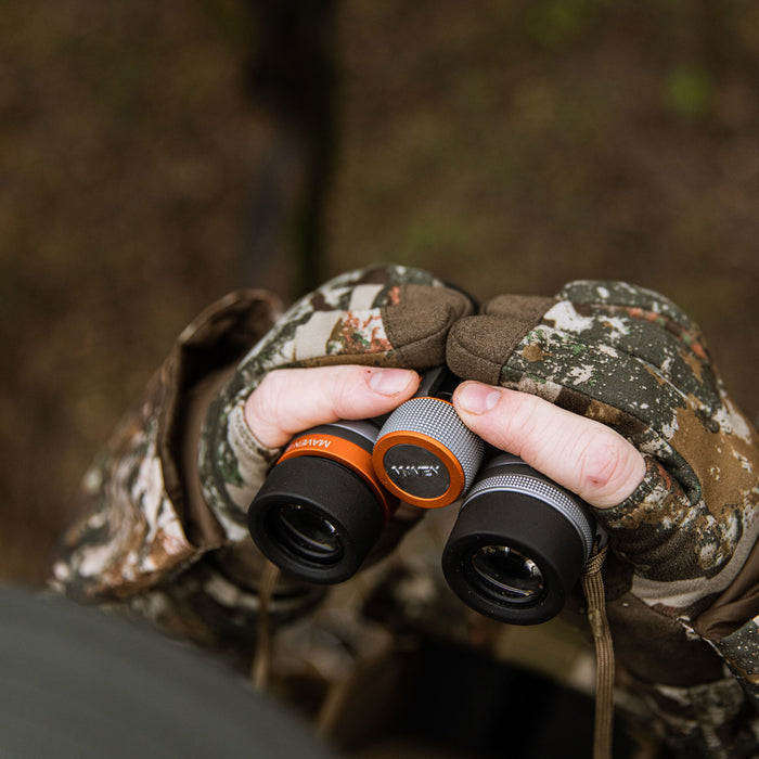 Hands in camouflage gloves hold black binoculars with orange rings, silver knurled focus rings, and a visible MAVEN logo.
