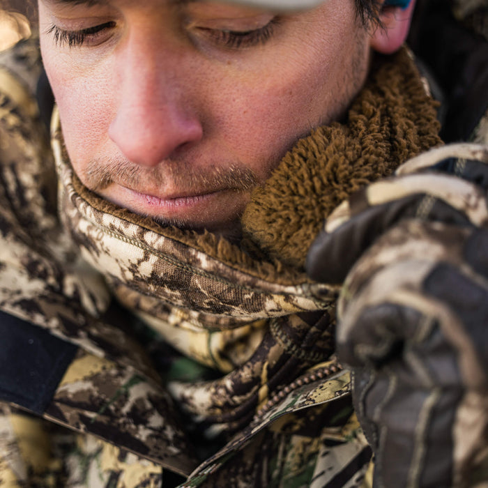Close-up of a person wearing an earth-toned camouflage jacket with a brown sherpa-lined collar, being adjusted by a gloved hand.