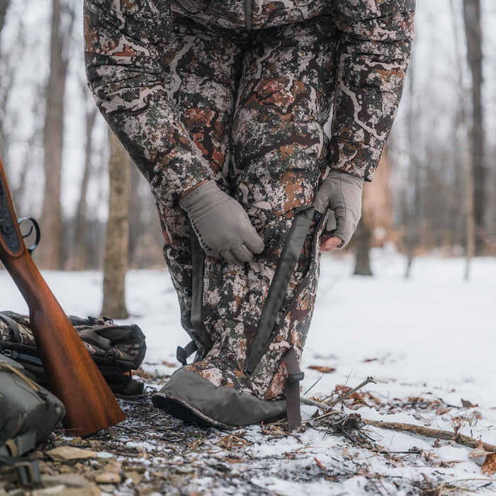 A person wearing a camouflage hunting jacket and pants with gray gloves is standing in a snowy forest, adjusting a black strap on the pants. A rifle and backpack are visible on the ground nearby.