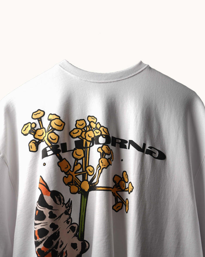 A close-up of a white crewneck sweatshirt featuring a graphic print of a yellow-orange flower with the word 'BLOOMING' and a spotted animal face in orange, black, and white.