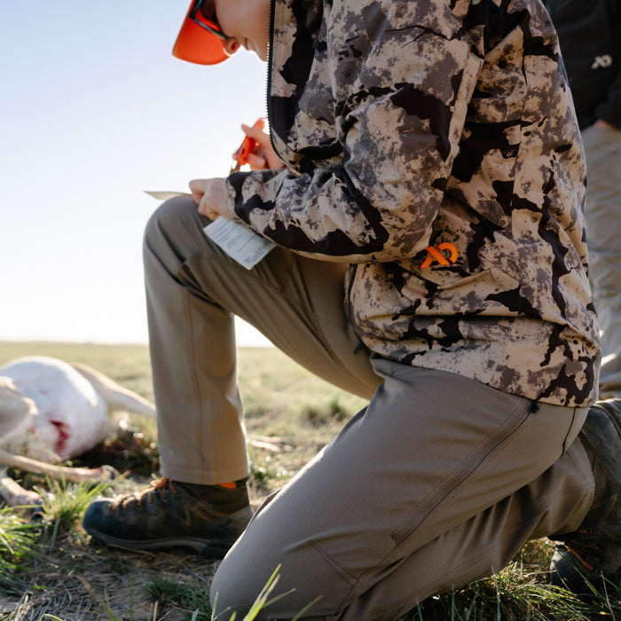 Hunter kneeling outdoors wearing a beige and dark brown camouflage jacket with an orange logo, olive green rugged pants with a vertical zippered thigh pocket, and dark brown hunting boots.