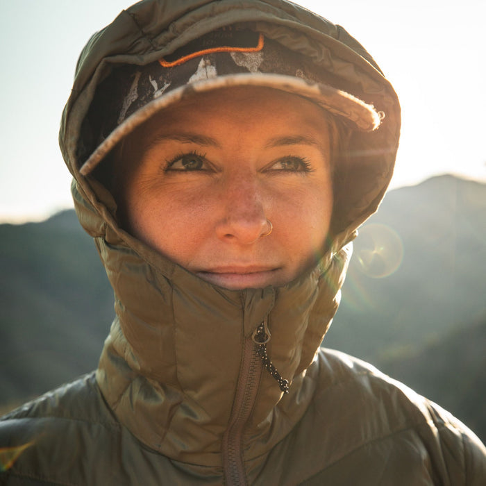 A woman wears an olive green hooded puffer jacket, zipped up, with a baseball cap visible under the hood.