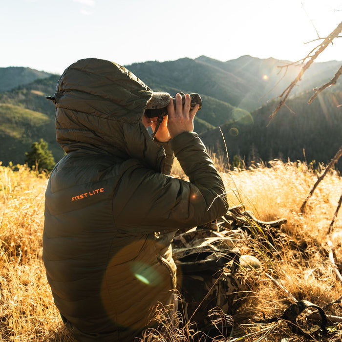 Olive green hooded puffy jacket with 'FIRST LITE' logo on the back, worn by a person looking through binoculars.