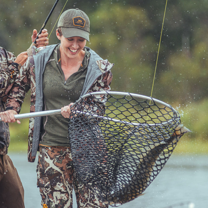 A smiling woman in a dark green cap with an orange logo, olive green quarter-zip shirt, and camouflage jacket and pants, holds a net with a fish splashing water.