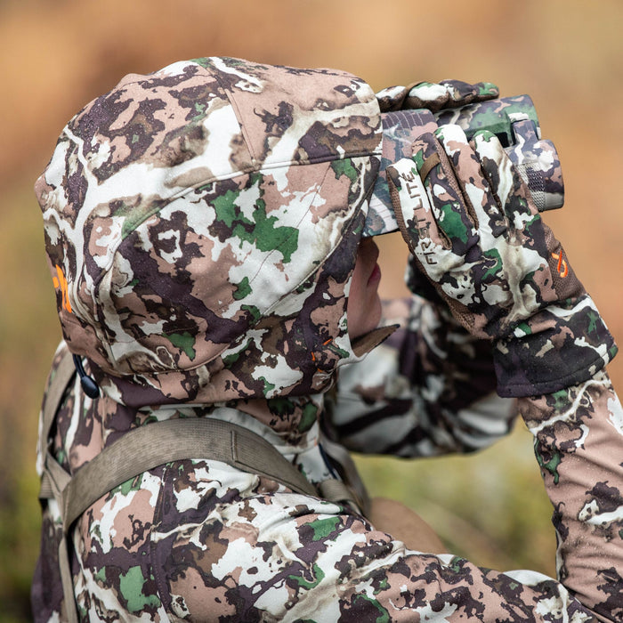 A person wears First Lite camouflage hunting apparel, including a hooded jacket, hat, and gloves with water droplets, while looking through matching binoculars.