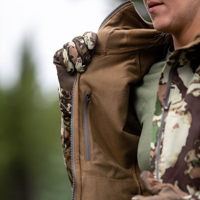 A person wears a brown jacket with camouflage sleeves, a main zipper, and a zippered chest pocket.