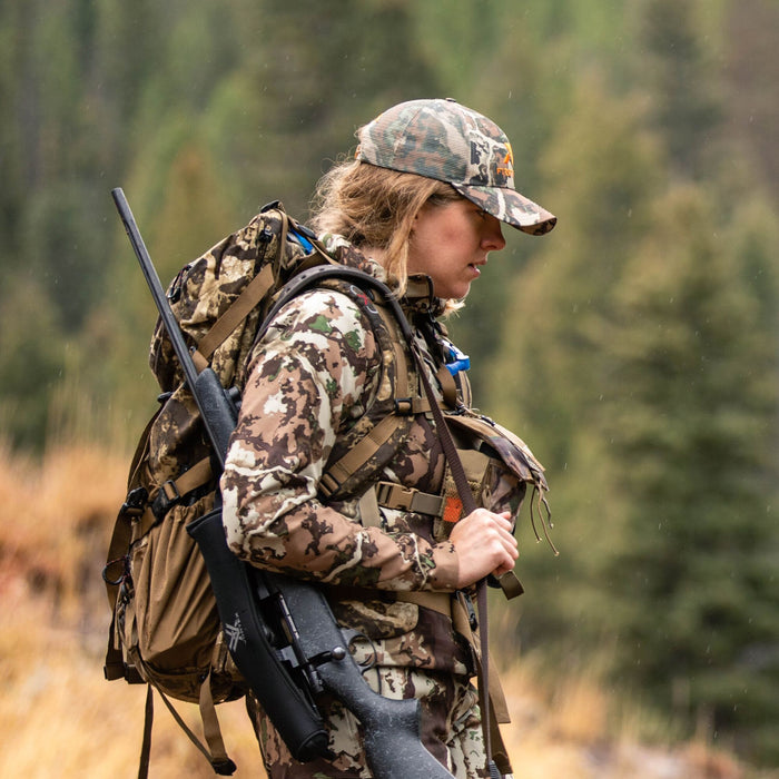 Woman in camouflage hunting jacket and hat with orange logo, carrying a tan backpack with a dark rifle attached, in a rainy forest.