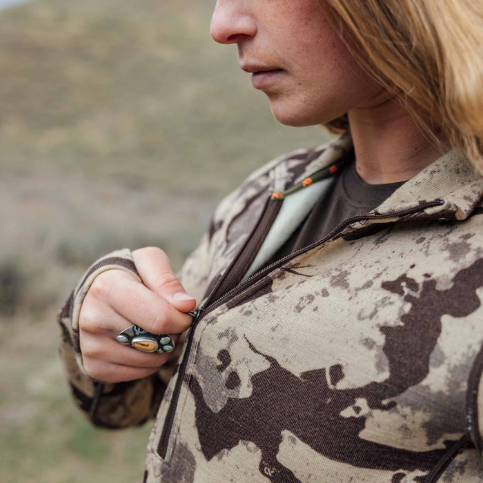 Close-up of a person zipping up a light beige and dark brown camouflage jacket, featuring a decorative ring and an orange zipper pull inside.