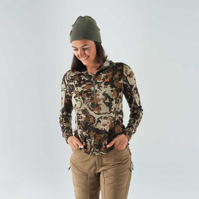 Woman modeling an olive green beanie, a camouflage 1/4-zip long-sleeve shirt with an orange logo, and tan outdoor pants.
