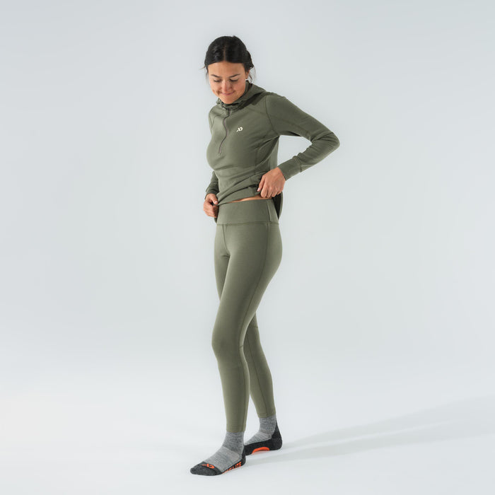 A woman wears an olive green base layer set, featuring a hooded quarter-zip top with a white chest logo and matching high-waisted leggings.