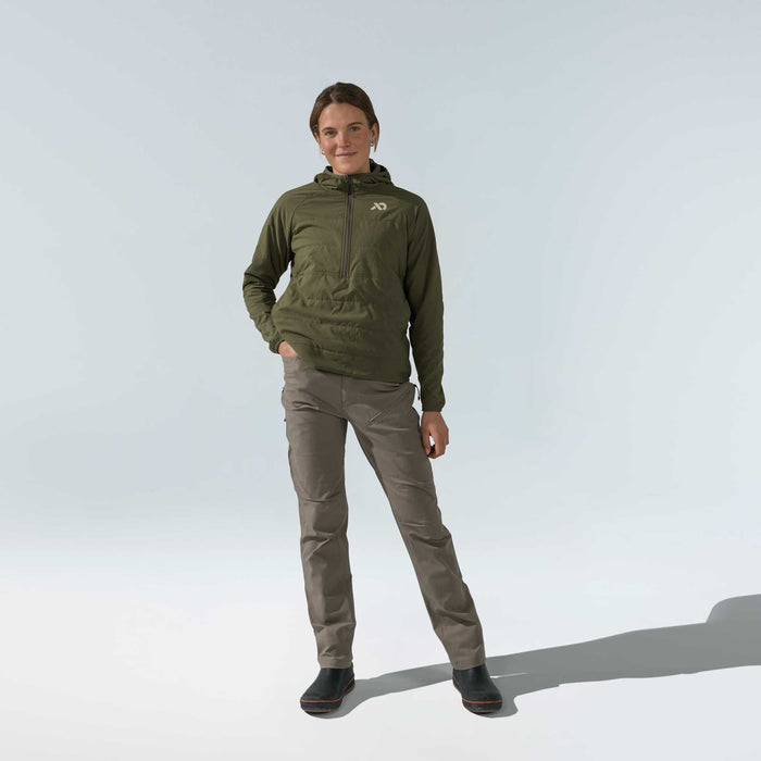 A woman wears an olive green hooded quarter-zip jacket with a chest logo and brown outdoor pants with knee articulation.