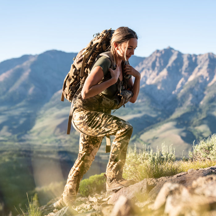 Woman wears tan camouflage pants and a large matching backpack while hiking a rocky mountain trail.