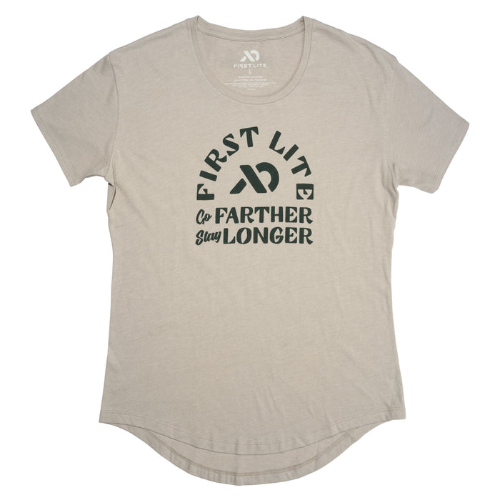 A light tan short-sleeve t-shirt with a crew neck, curved hem, and a dark green "FIRST LITE Go FARTHER Stay LONGER" graphic on the front.