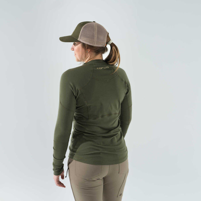 Rear view of a woman wearing an olive green First Lite long-sleeve shirt with a gold logo, a green and tan mesh trucker hat, and tan pants.