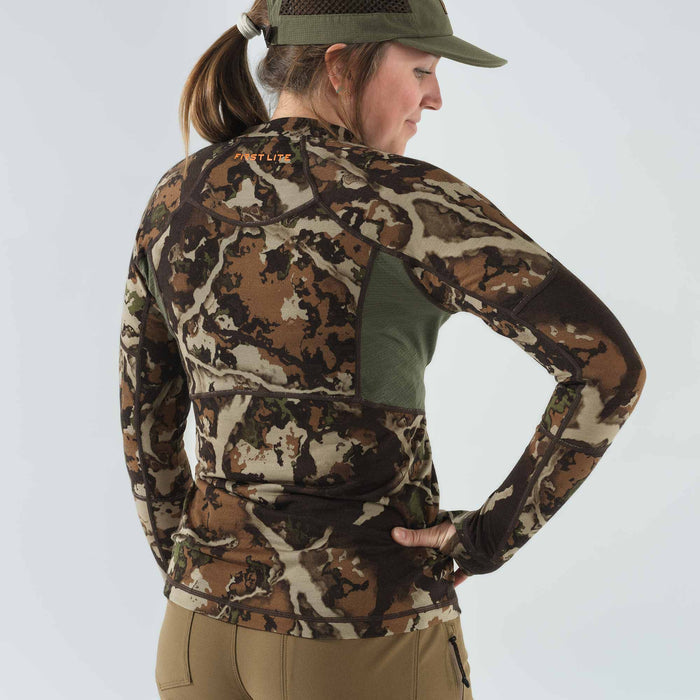 A woman models a brown, tan, and green camouflage long-sleeve base layer shirt featuring green mesh panels and an orange "FIRST LITE" logo on the upper back.