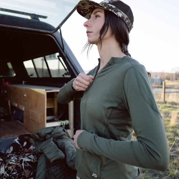 Woman zipping up a dark olive green quarter-zip long-sleeve shirt with a camouflage hat.